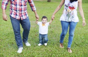 Garoto pequeno com síndrome de Down aparece caminhando no centro da foto. A mãe e o pai estão um de cada lado dele segurando a sua mãe e ajudando o pequeno a caminhar.