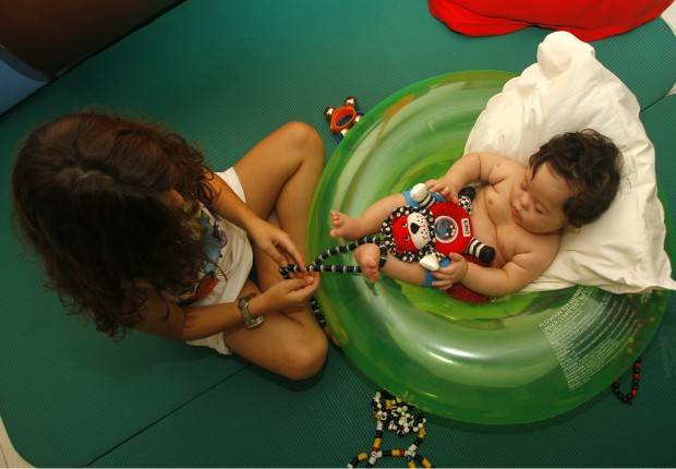 Um bebê de pele branca com síndrome de Down está deitado no centro de uma boa de piscina em forma de rosca. Ele está apoiado em um travesseiro e segura um bichinho de pelúcia. Uma garota morena sentada à frente do bebê segura uma corda e puxa o pé dele para fazer exercícios de estimulação precoce.