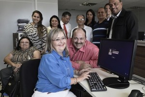 Mulher com síndrome de Down sentada em mesa com coputador, sorridente, com colegas de trabalho à volta.