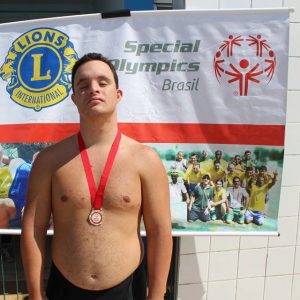 Pedro Fernandes tem síndrome de Down e é um campeão na natação. Ele aparece na foto com calção de natação e medalha de outro no peito. Atrás tem um painel escrito Special Olympics Brasil e com algumas fotos de atletas brasileiros com deficiência intelectual. O sonho de Pedro é poder inspirar o mundo com a sua história.