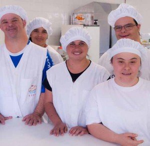 Um grupo de jovens com e sem síndrome de Down posa para a foto em seu local de trabalho. Eles usam um uniforme branco e uma touca branca na cabeça. Todos sorriem. A imagem exemplifica a importância da autonomia e inclusão social de pessoas com deficiência.