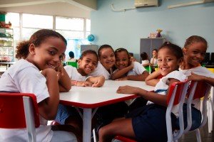 Alunas do ensino público estão sentadas em volta de uma carteira na sala de aula. São cinco garotas, todas uniformizadas e negras. A garota no centro tem síndrome de Down, enquanto as outras não apresentam nenhuma deficiência. A foto ilustra o tema da educação inclusiva.